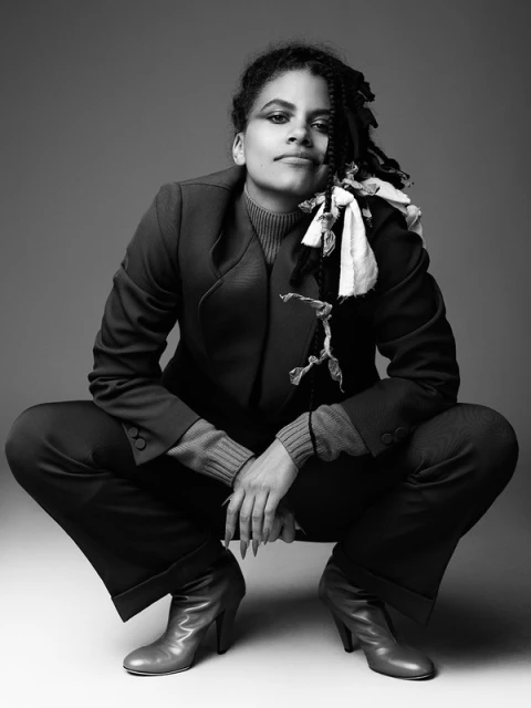 Portrait of a woman with dark hair and a blazer crouches, looking at the camera. She wears a blazer, sweater, pants, and heeled boots.