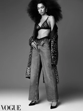 Model with voluminous afro hair wearing a leopard print coat over black lingerie and wide-leg jeans in a studio portrait.