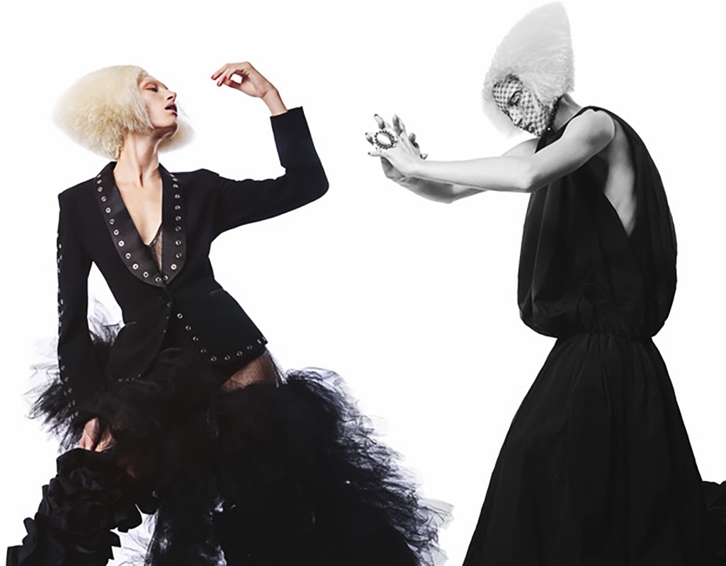 Two fashion models in avant-garde black outfits and sculptural white hair, posing against a stark white backdrop in a photo studio.
