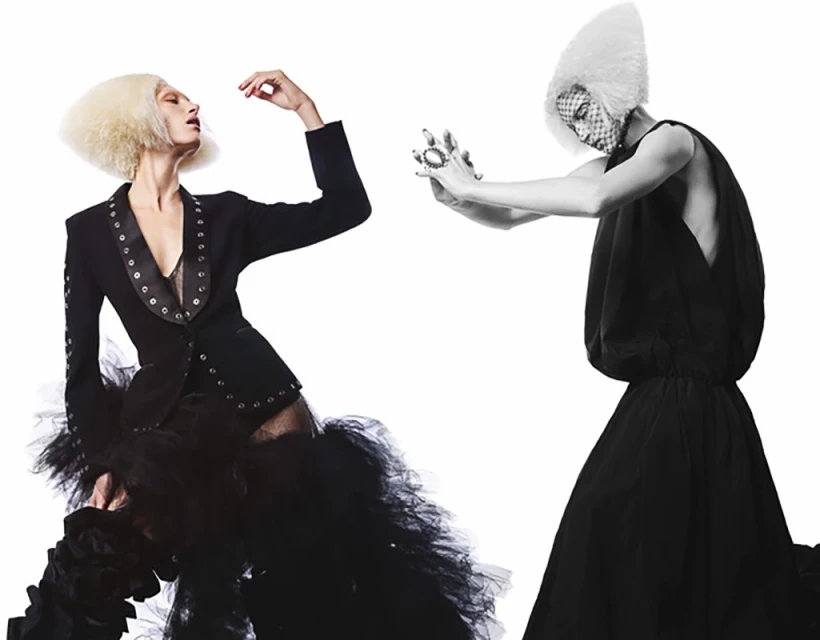 Two fashion models in avant-garde black outfits and sculptural white hair, posing against a stark white backdrop in a photo studio.