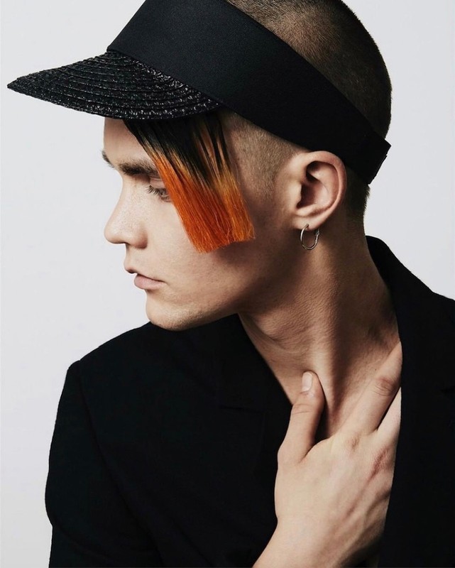 Close-up profile portrait of a young adult male wearing a black visor and blazer, orange-colored fringe, silver hoop earrings, studio setting.