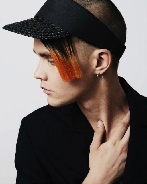 Close-up profile portrait of a young adult male wearing a black visor and blazer, orange-colored fringe, silver hoop earrings, studio setting.