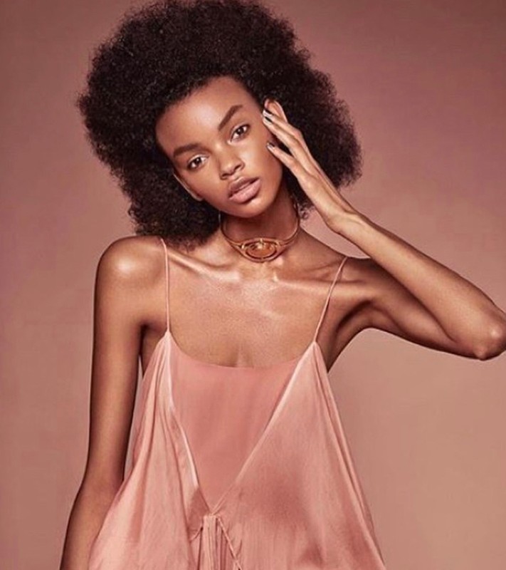 Close-up portrait of a Black woman with an afro hairstyle wearing a slip dress and a metallic choker, against a monochromatic warm background.
