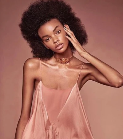 Close-up portrait of a Black woman with an afro hairstyle wearing a slip dress and a metallic choker, against a monochromatic warm background.