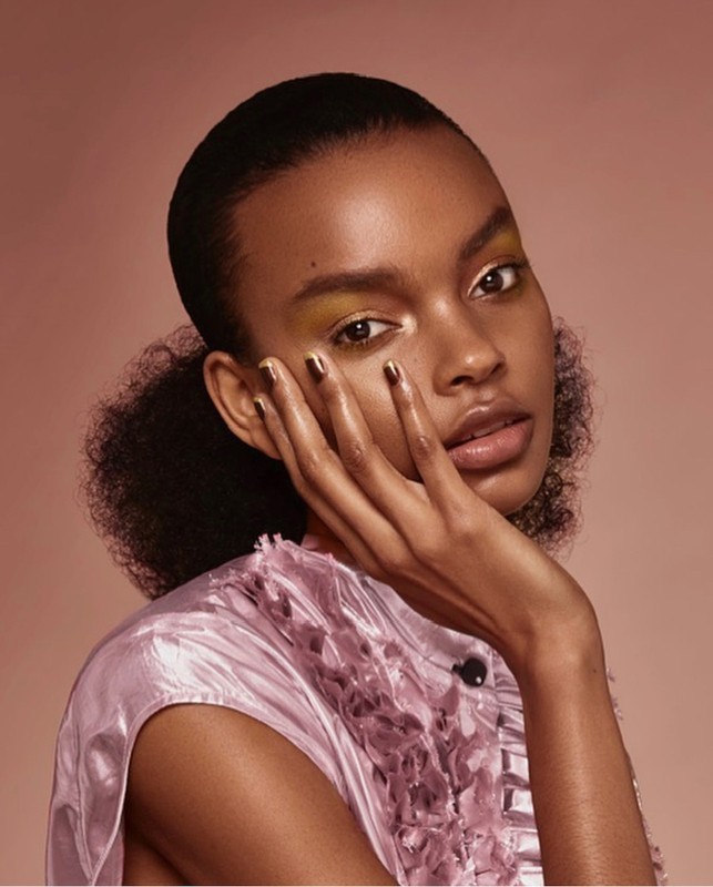Close-up studio shot of an African American woman with her hand to her face, wearing a pink frilled top and eyeshadow makeup.