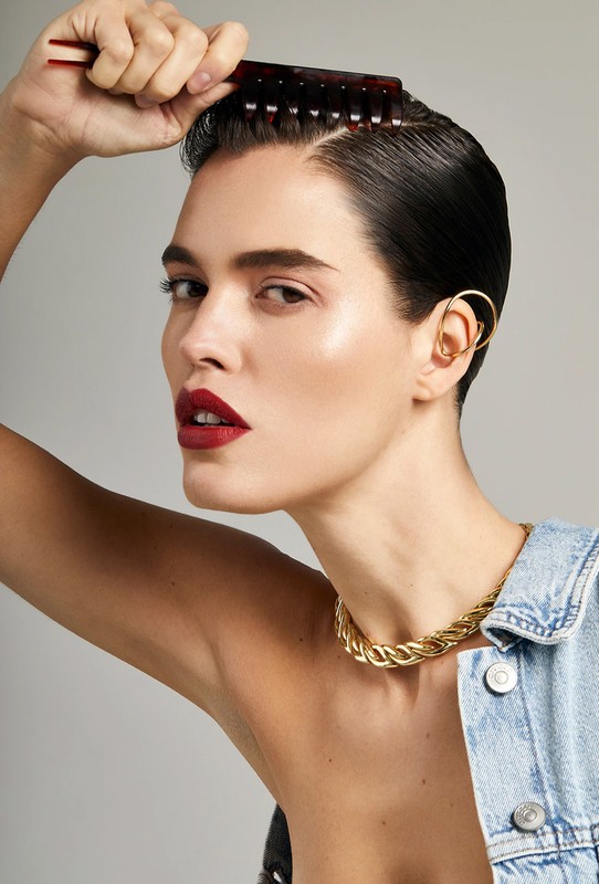 Portrait of a woman combing her hair wearing a gold chain necklace, denim jacket, and gold ear cuff against a grey background.