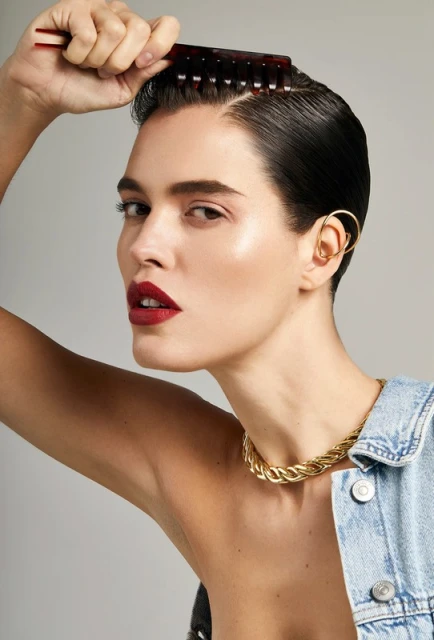 Portrait of a woman combing her hair wearing a gold chain necklace, denim jacket, and gold ear cuff against a grey background.
