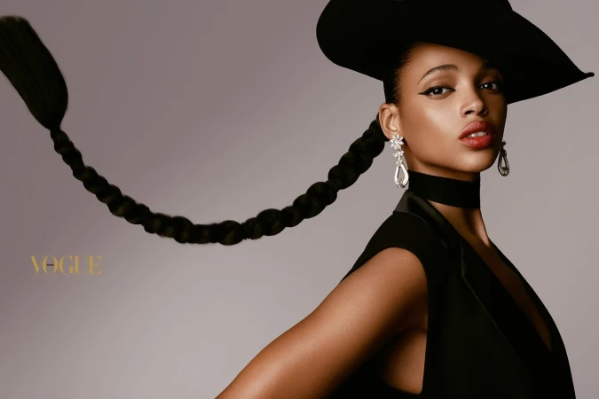 Black woman wearing a wide-brimmed black hat, statement earrings, and a black top against a grey seamless background
