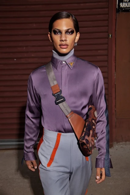 Portrait of a person with dramatic eye makeup wearing a lilac satin shirt, grey pants with orange accents, and a brown crossbody bag.