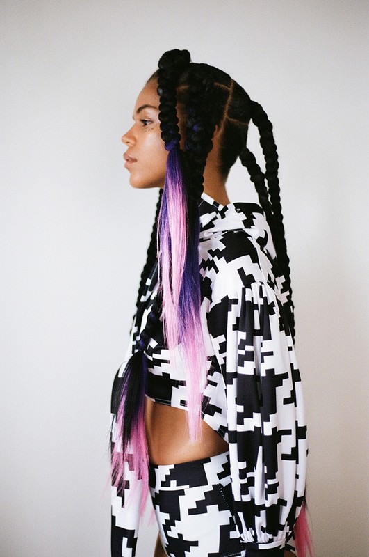 Profile view of a woman with long braids and a black-and-white patterned two-piece outfit standing against a neutral backdrop.