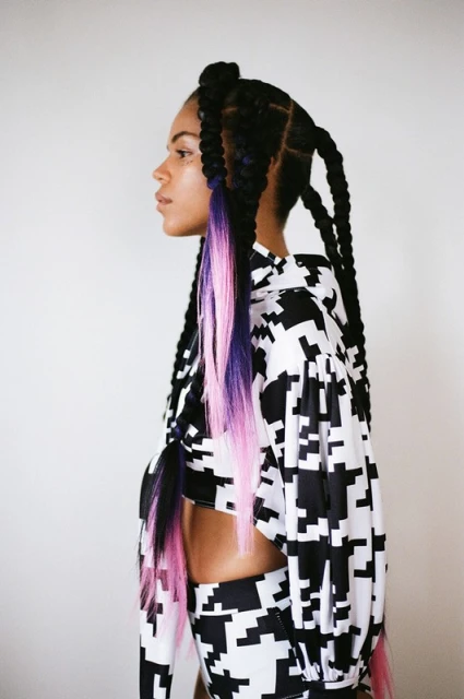 Profile view of a woman with long braids and a black-and-white patterned two-piece outfit standing against a neutral backdrop.