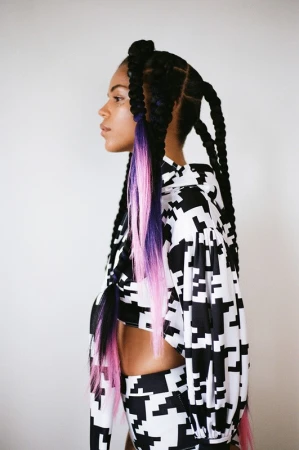 Profile view of a woman with long braids and a black-and-white patterned two-piece outfit standing against a neutral backdrop.