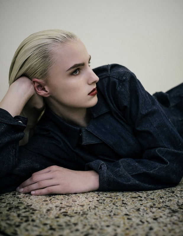 Blonde woman in denim laying indoors. Close-up portrait showing a person in a blue jacket and red lipstick on a textured floor.
