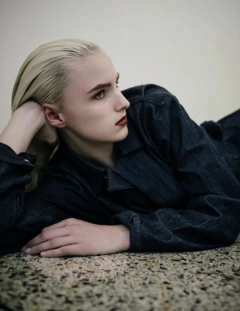 Blonde woman in denim laying indoors. Close-up portrait showing a person in a blue jacket and red lipstick on a textured floor.