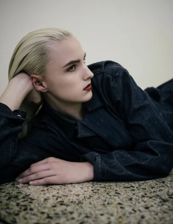 Blonde woman in denim laying indoors. Close-up portrait showing a person in a blue jacket and red lipstick on a textured floor.