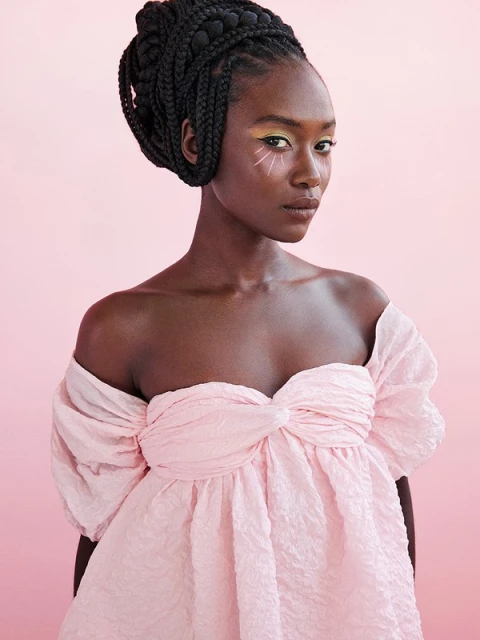Elegant portrait of a Black woman with braided hair and stylized makeup, wearing a pink dress against a light pink backdrop.