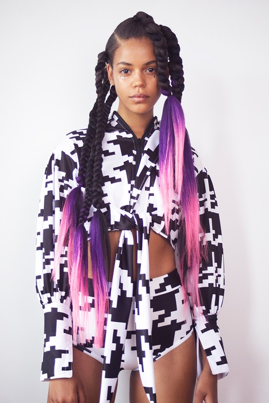A young woman poses in a matching houndstooth crop top and shorts, showcasing her black braids with pink and purple dyed ends in a studio.