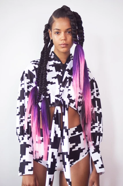 A young woman poses in a matching houndstooth crop top and shorts, showcasing her black braids with pink and purple dyed ends in a studio.