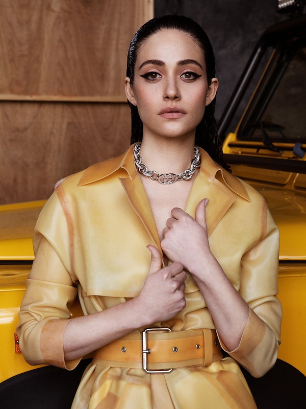Emmy Rossum poses in a yellow vinyl coat and silver necklace in an indoor setting with a yellow car partially visible behind her.