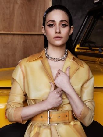Emmy Rossum poses in a yellow vinyl coat and silver necklace in an indoor setting with a yellow car partially visible behind her.