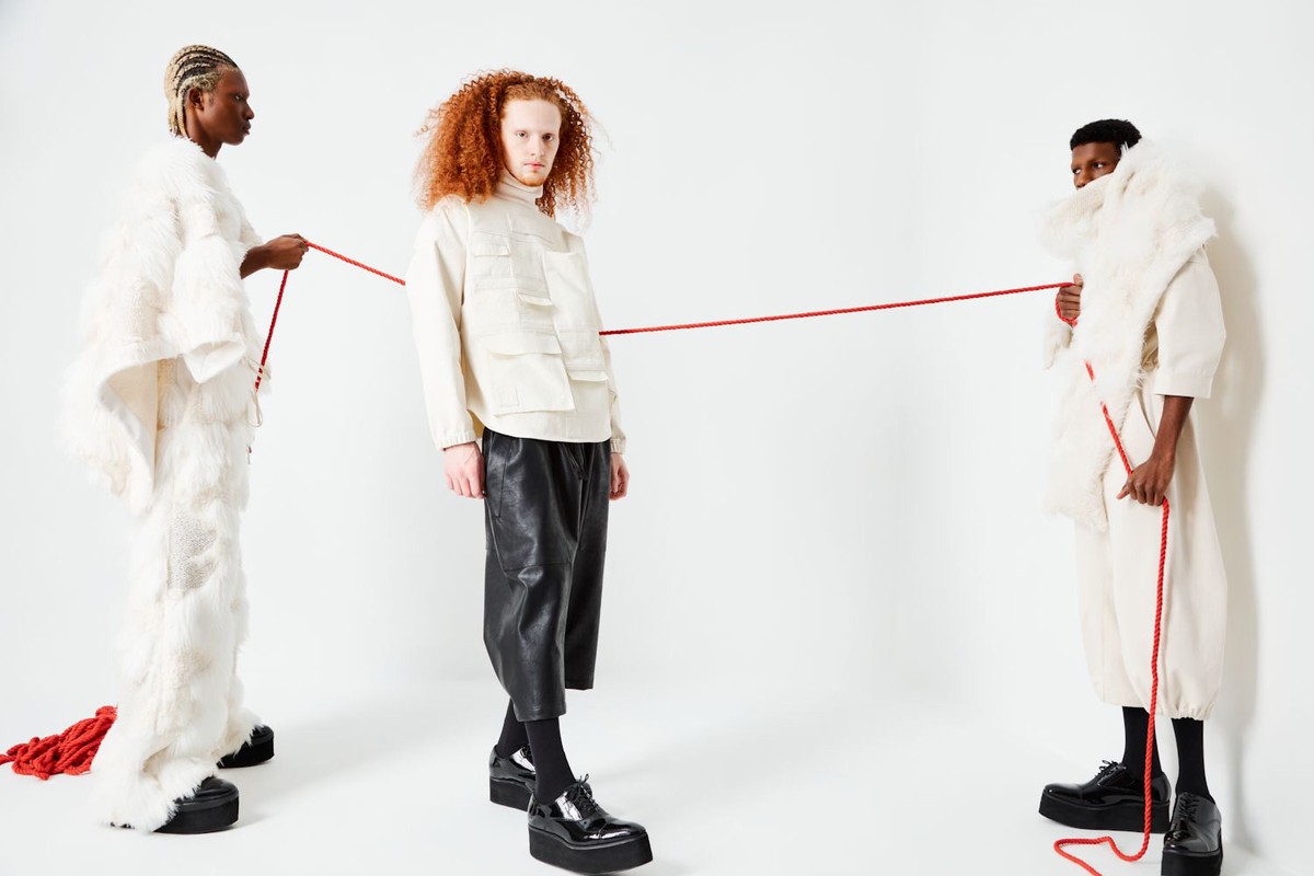 Three fashion models in avant-garde white and black clothing pose with a red string against a clean white backdrop in a studio setting.