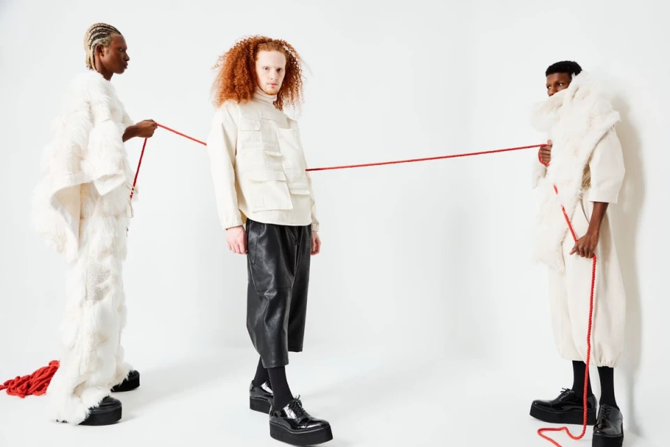 Three fashion models in avant-garde white and black clothing pose with a red string against a clean white backdrop in a studio setting.