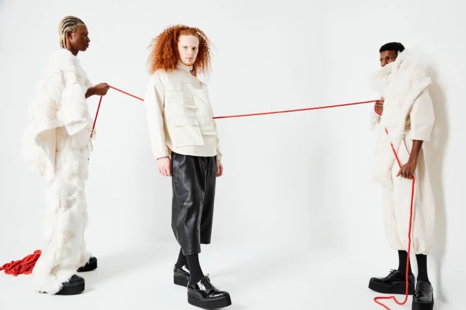 Three fashion models in avant-garde white and black clothing pose with a red string against a clean white backdrop in a studio setting.
