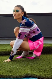 Model crouching outdoors in a star pattern sweater, hot pink mini skirt, and metallic pink pointed kitten heels on artificial turf.