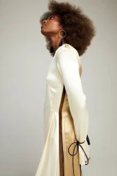 Model with voluminous afro hairstyle wearing a cream satin dress with brown panels and hoop earrings against a neutral background.
