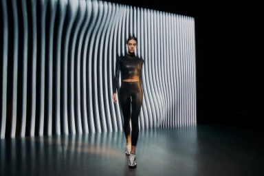 Model in black athletic apparel and sneakers walking toward camera against a curved vertical slat backdrop.