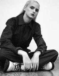 Monochrome portrait of a seated model wearing a dark utility jumpsuit and light boots against a plain wall.