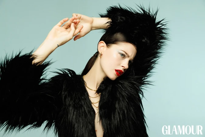 Woman in black faux fur jacket with red lipstick and gold chains posing against a solid turquoise background