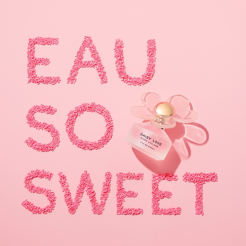 Close-up of a Daisy Love Marc Jacobs perfume bottle with Eau So Sweet written in pink sprinkles on a light pink background.