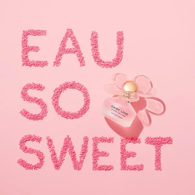 Close-up of a Daisy Love Marc Jacobs perfume bottle with Eau So Sweet written in pink sprinkles on a light pink background.