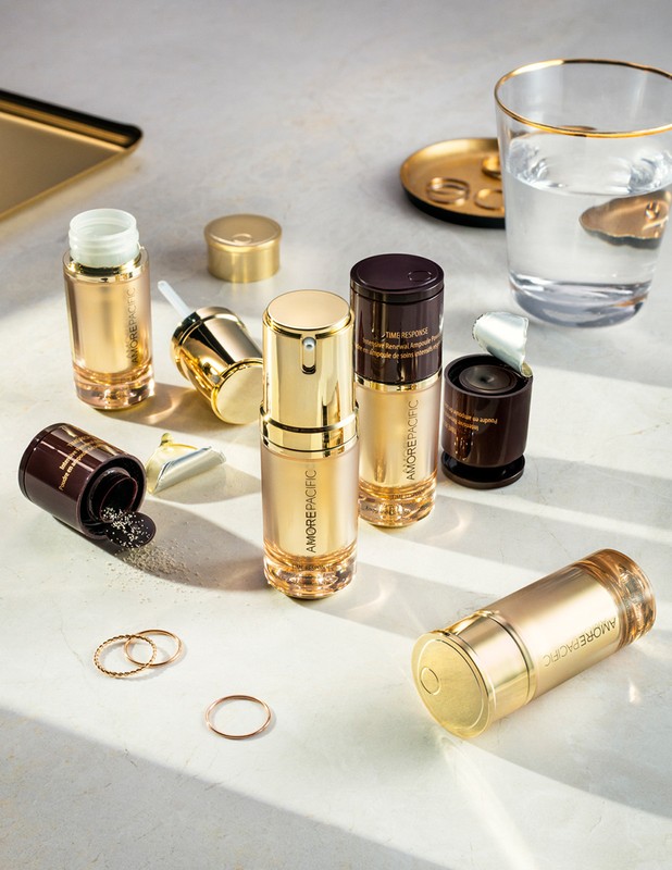 A skincare product flat lay featuring golden-toned AMOREPACIFIC bottles, containers with dark brown accents, and gold accessories on a light surface.