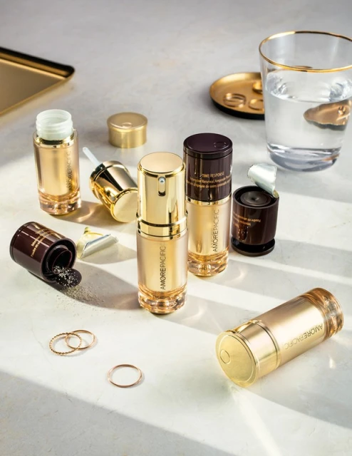 A skincare product flat lay featuring golden-toned AMOREPACIFIC bottles, containers with dark brown accents, and gold accessories on a light surface.