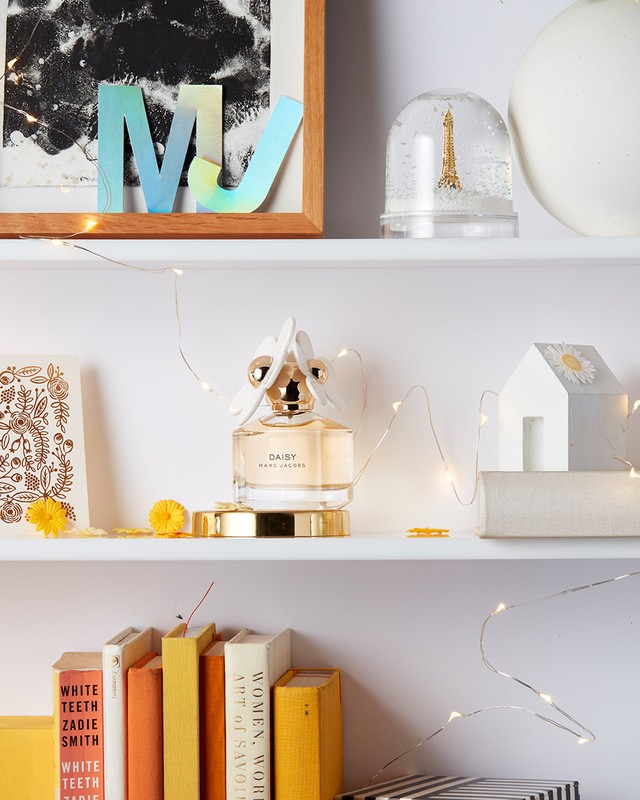 Daisy Marc Jacobs perfume bottle on a white shelf with books, decorations, and fairy lights creating a soft, inviting aesthetic.