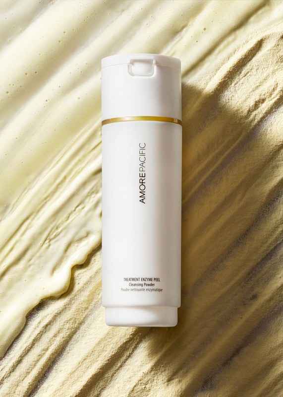 Amore Pacific skincare product, a white cylindrical bottle with gold accent, stands on a textured cream-colored background in a close-up shot.