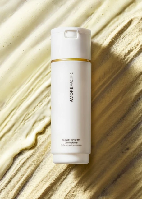 Amore Pacific skincare product, a white cylindrical bottle with gold accent, stands on a textured cream-colored background in a close-up shot.