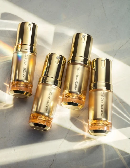 Four gold-toned AmorePacific skincare bottles are arranged diagonally on a textured, light-grey surface with soft shadows and rainbow reflections.