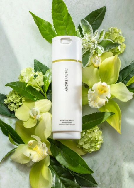 White Amorepacific bottle surrounded by green leaves and white orchids on a light marble background in a beauty product shot.