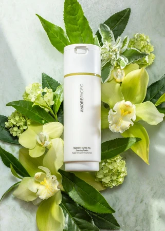 White Amorepacific bottle surrounded by green leaves and white orchids on a light marble background in a beauty product shot.