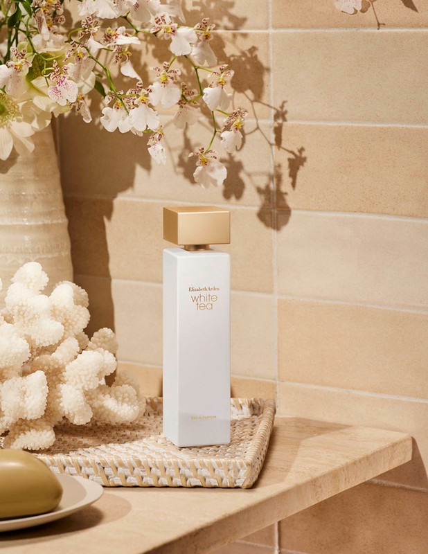 Still life shot of Elizabeth Arden White Tea perfume bottle with white flowers, coral, and soap on a tiled countertop.