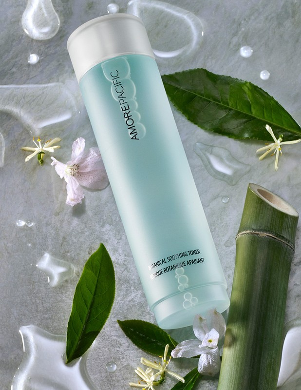 Overhead shot of AMOREPACIFIC botanical soothing toner surrounded by green leaves, white flowers, bamboo, and water droplets on grey stone.