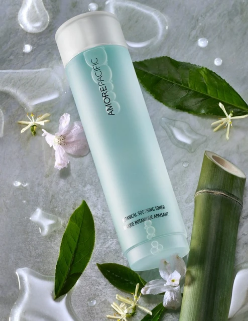 Overhead shot of AMOREPACIFIC botanical soothing toner surrounded by green leaves, white flowers, bamboo, and water droplets on grey stone.