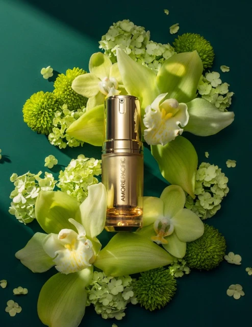 Golden skincare serum bottle by Amore Pacific on green backdrop surrounded by blooming white orchids and green hydrangeas.