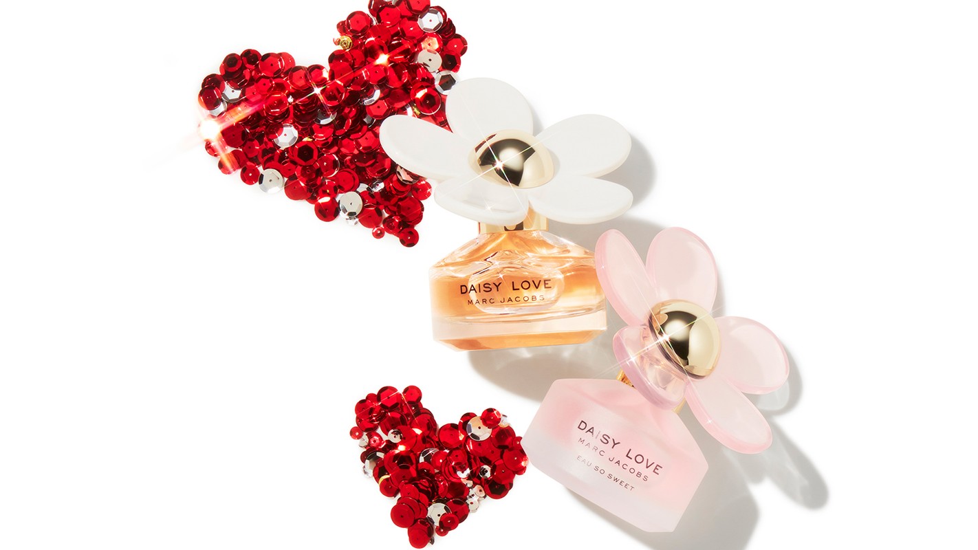 Daisy Love Marc Jacobs fragrance bottles with floral caps surrounded by red and silver sequin hearts on a white backdrop.