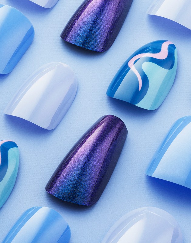 Overhead studio shot of a set of artificial nails in various colors, including shades of blue, purple, and white, arranged on a light blue surface.