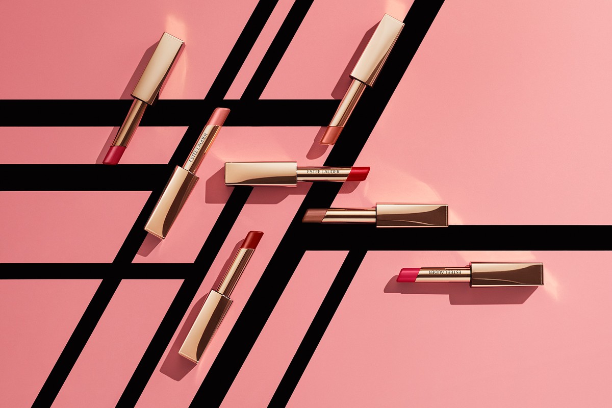 Several Estee Lauder lipsticks are arranged on a pink surface with black stripes. The lipsticks are gold with varied color lipstick shades.