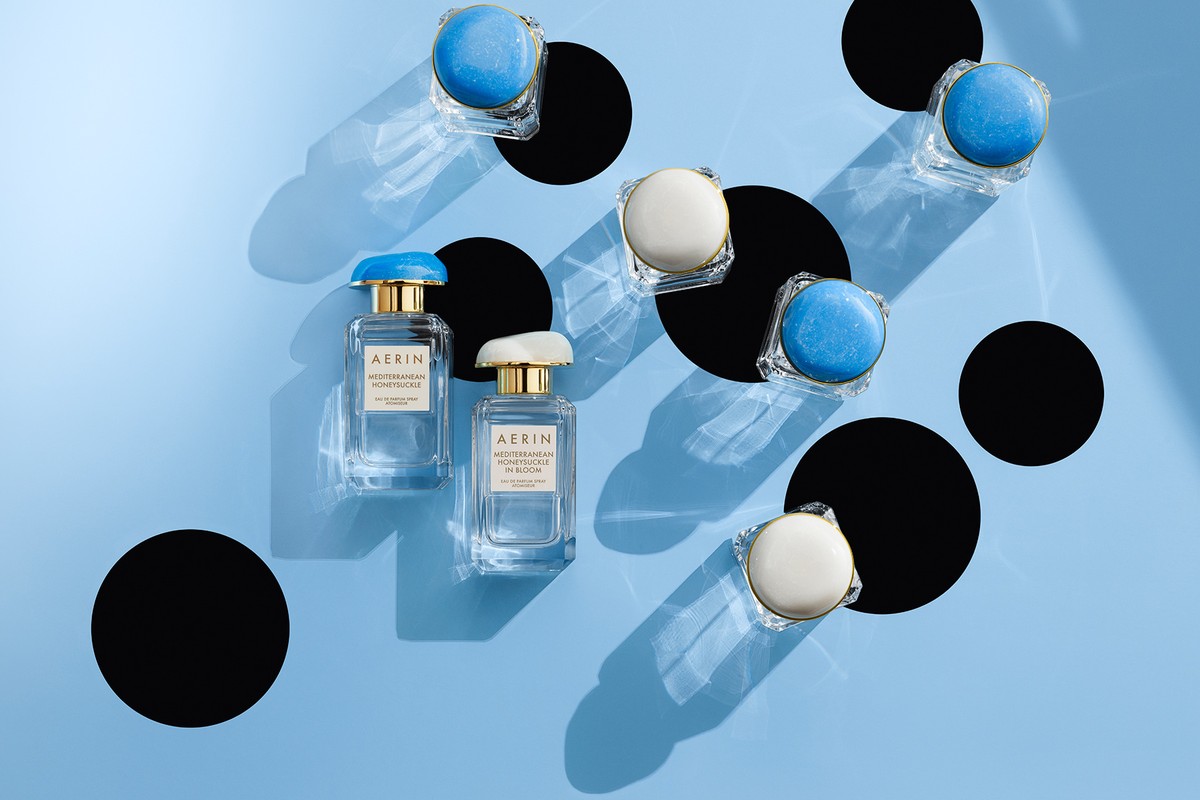 Two AERIN perfume bottles and several solid perfume compacts scattered on a light blue background, with black geometric shapes adding contrast.
